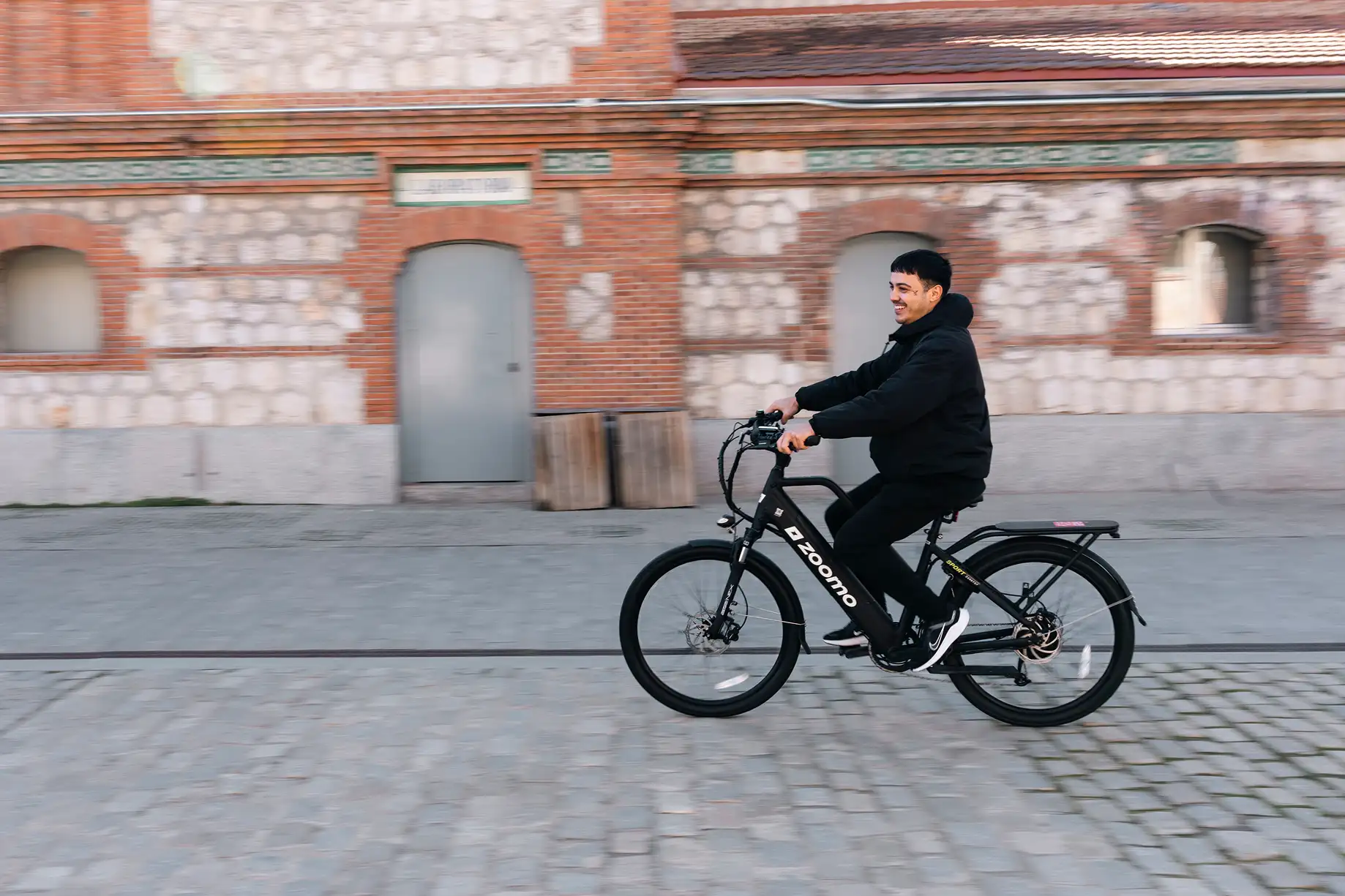 Man wearing black jacket riding a black Zoomo electric bike on a cobblestone street in front of a brick building.