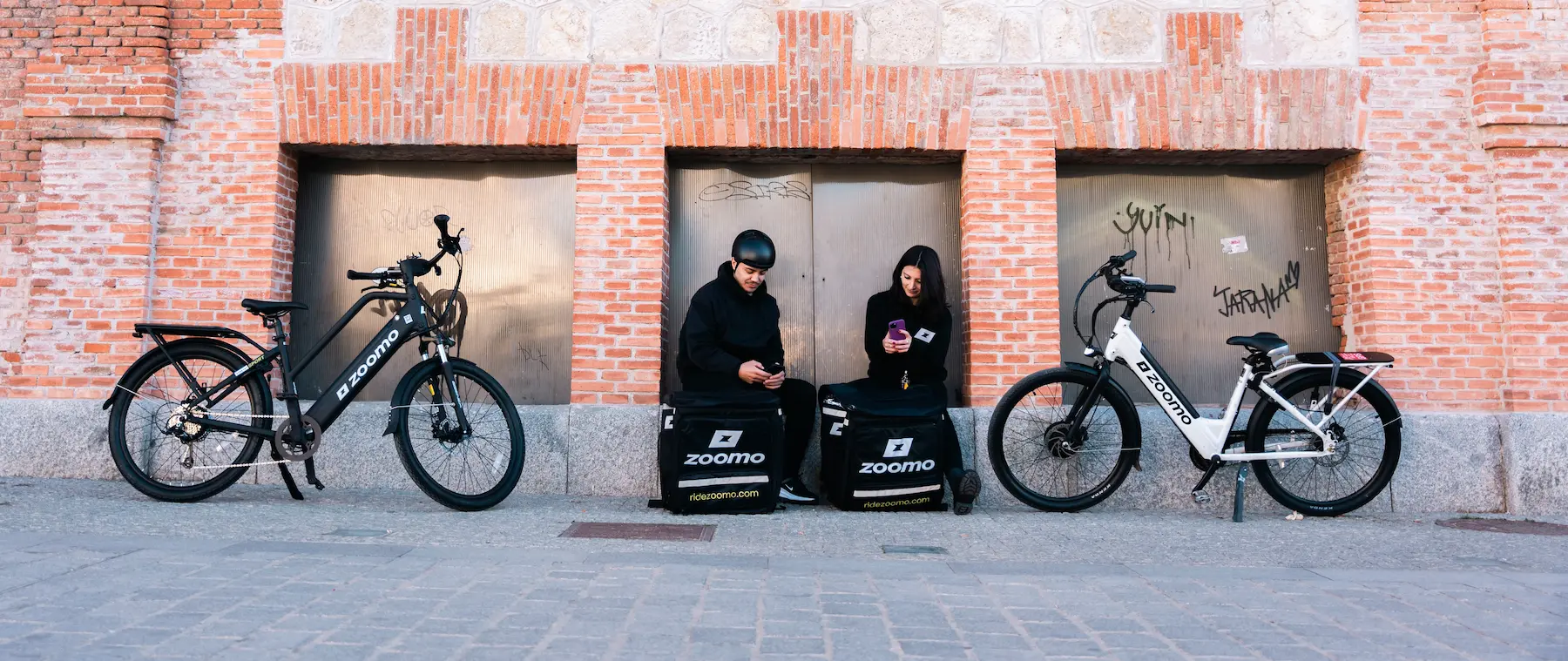 Two people sitting on Zoomo delivery bags between black and white Zoomo bikes by a brick wall.