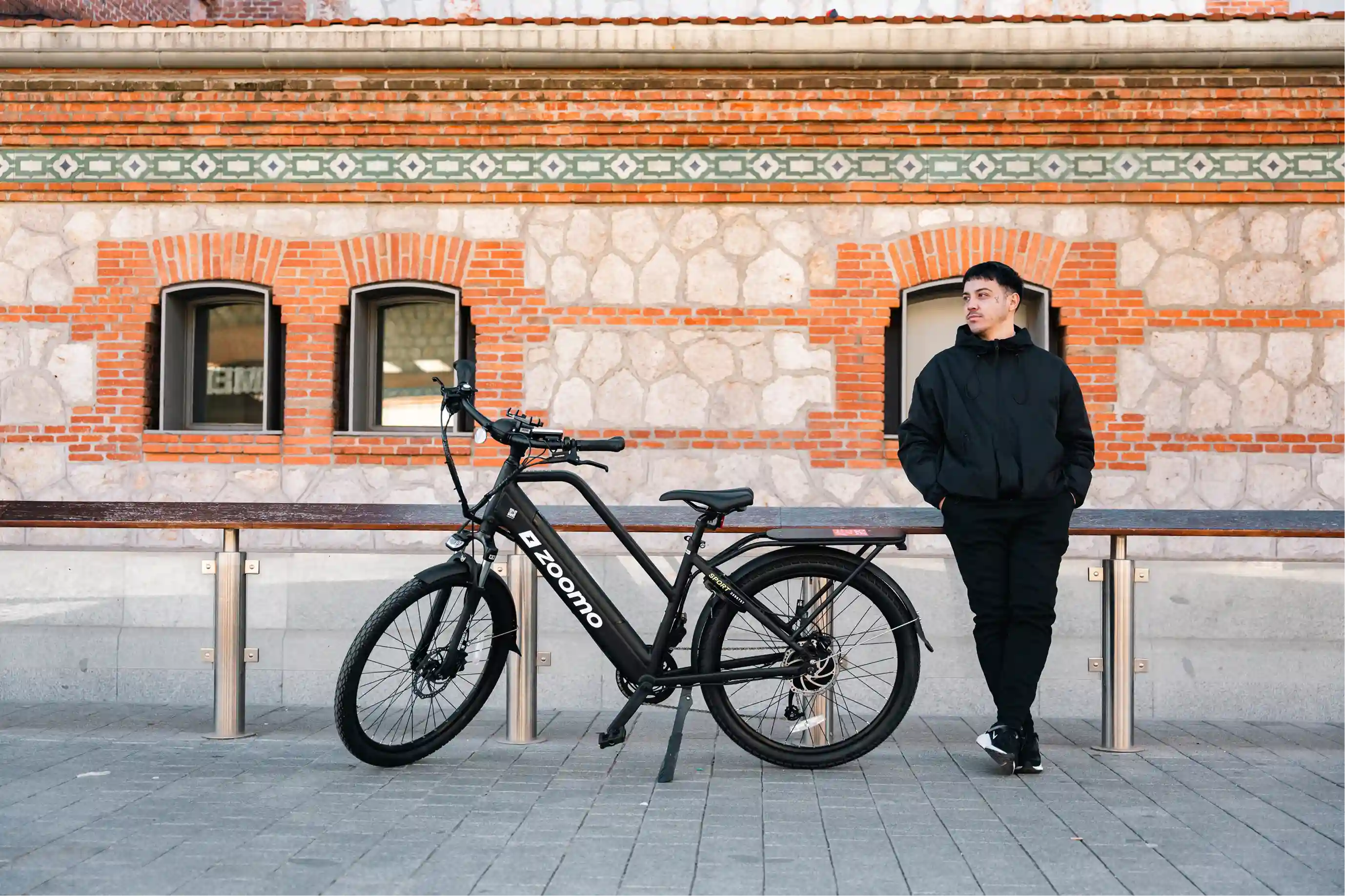 Man in black jacket and pants standing next to a black Zoomo electric bike against a brick and stone wall.