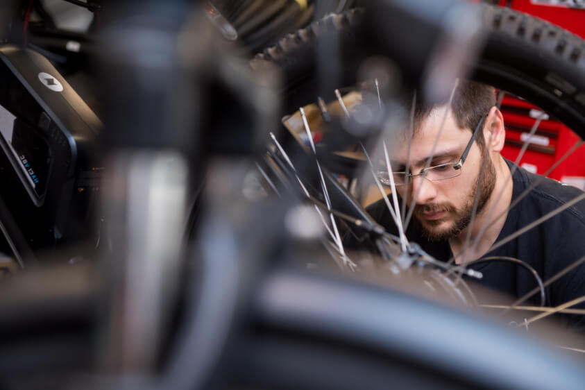 Man with glasses focused on repairing a bicycle wheel in a workshop.