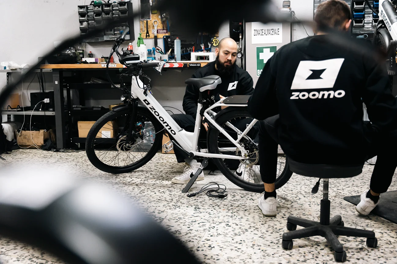 Two technicians in Zoomo jackets working on a white Zoomo electric bike inside a workshop.