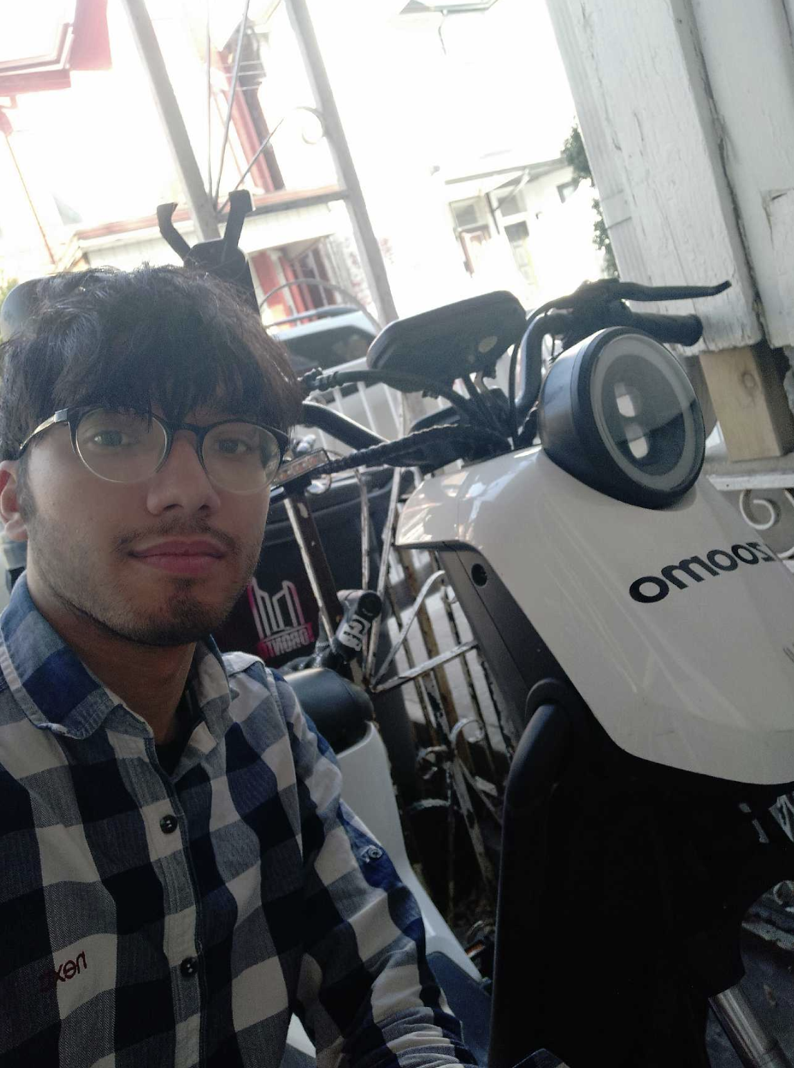 Young man with glasses and checkered shirt beside a white OMO scooter parked near a wooden wall.