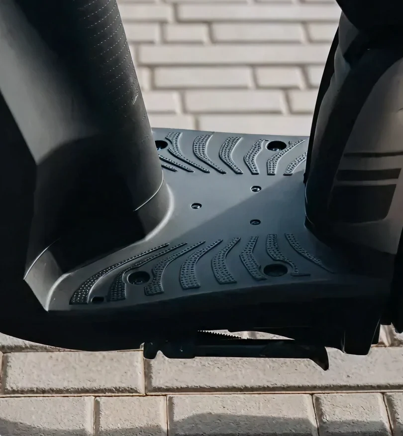 Close-up of the footrest area of a black electric scooter on a brick pavement.
