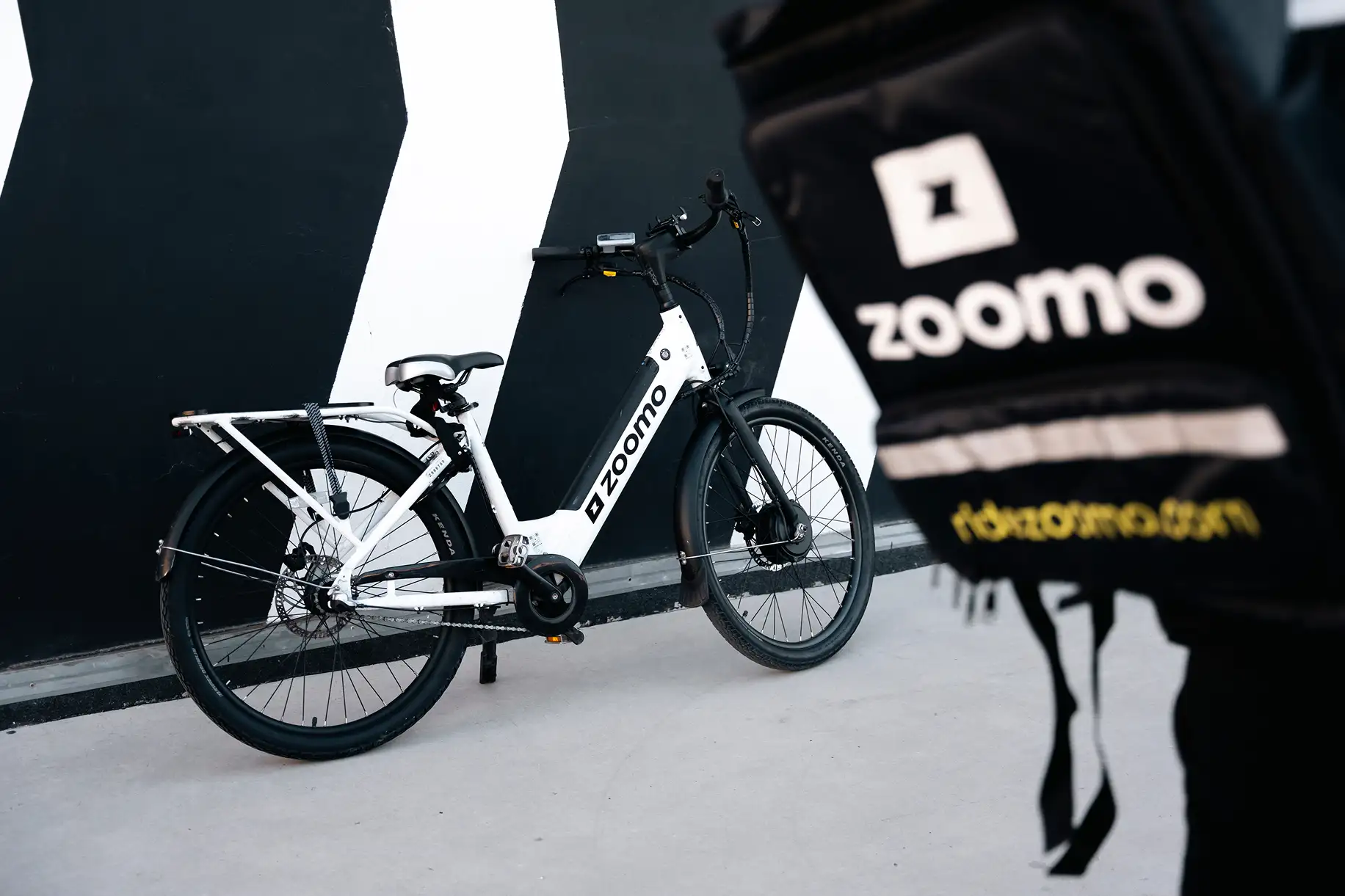 White electric bicycle with black details branded 'Zoomo' parked against a black and white striped wall.