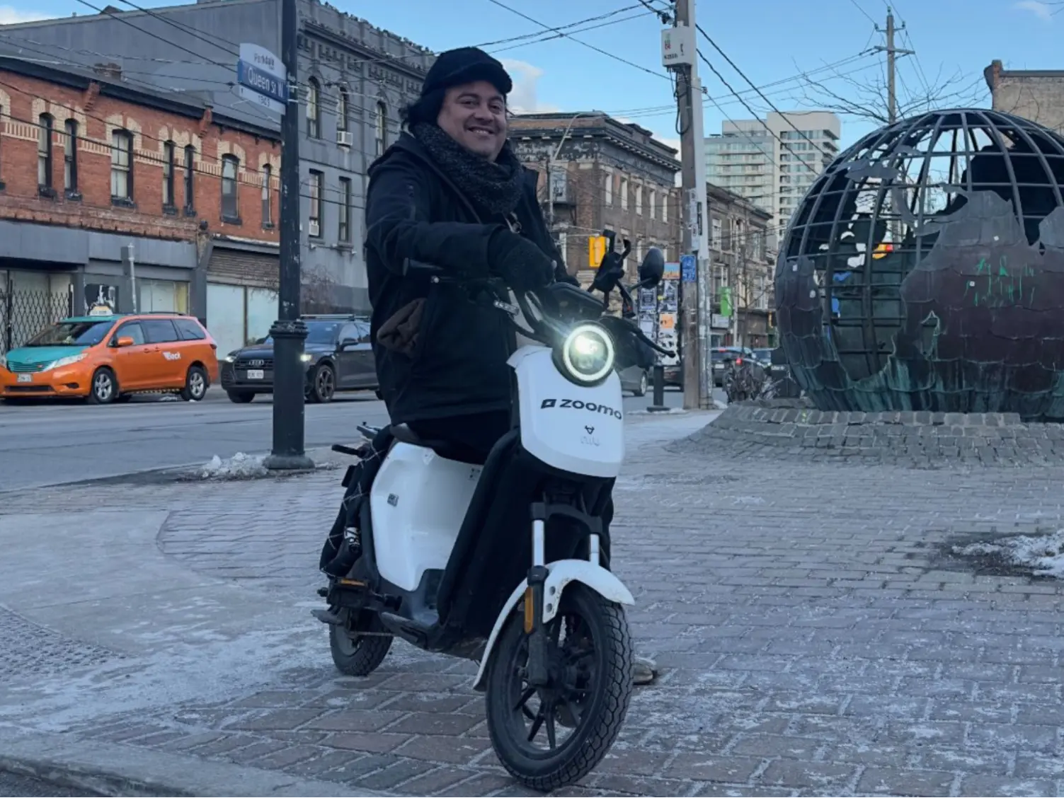 Person smiling on a white Zoomo electric scooter near a globe sculpture on a city sidewalk.