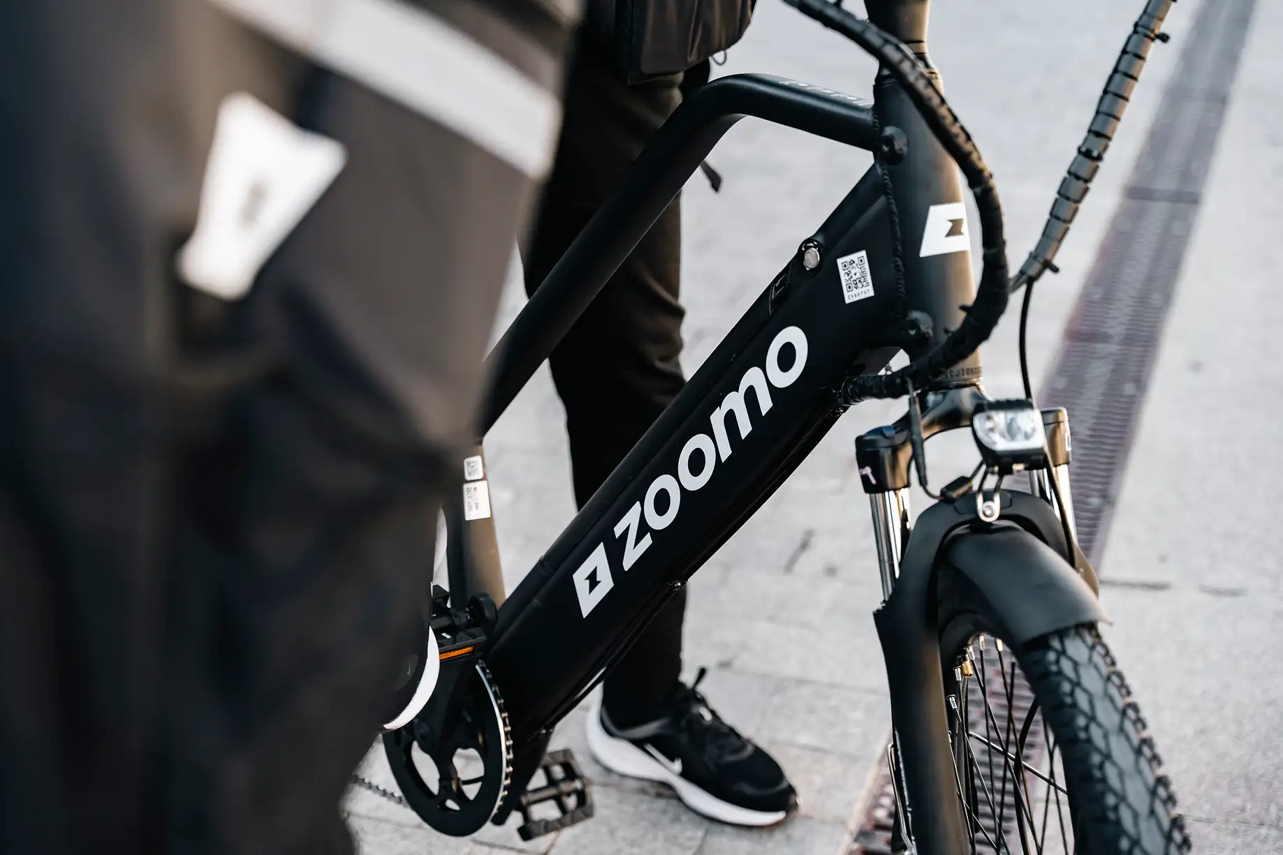 Close-up of a black Zoomo electric bike with rider's legs and black sneakers visible on a paved street.