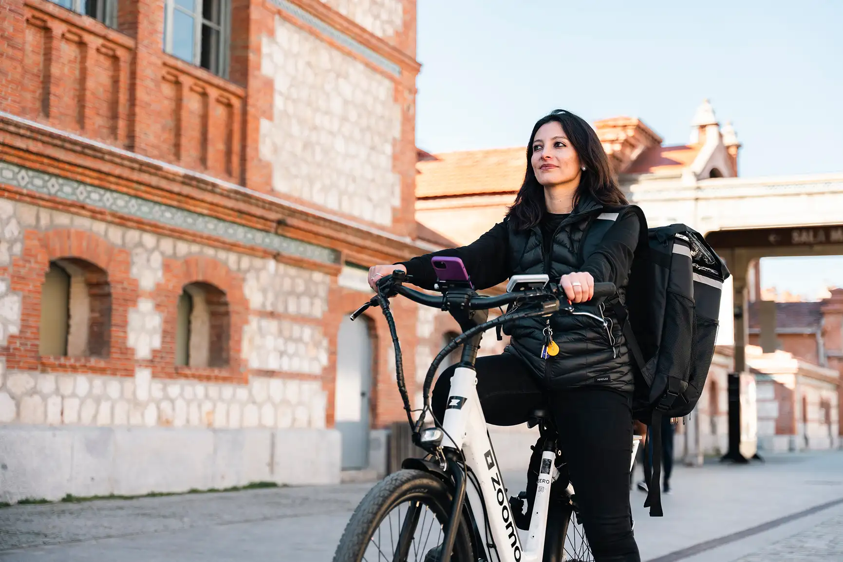 Woman in black jacket sitting on a white Zoomo electric bike with a large black backpack, in front of a brick and stone building.