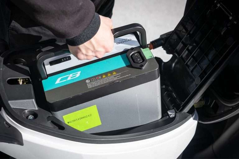 A hand lifting a large rectangular electric vehicle battery from a compartment with a black handle on top.